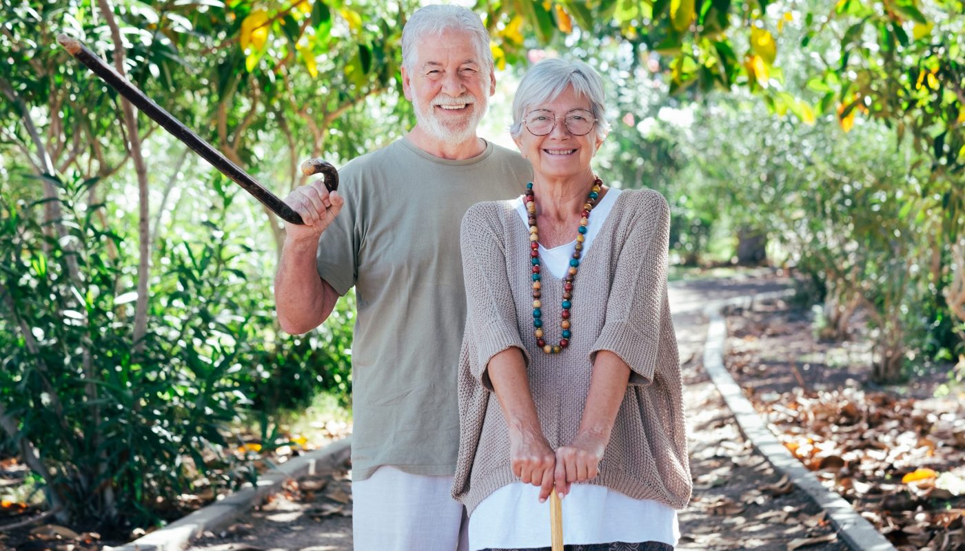 Beautiful white-haired senior couple walking in the woods with h Beautiful white-haired senior couple walking in the woods with help of a walking cane. Smiling elderly grandparents enjoy healthy lifestyle in public park
